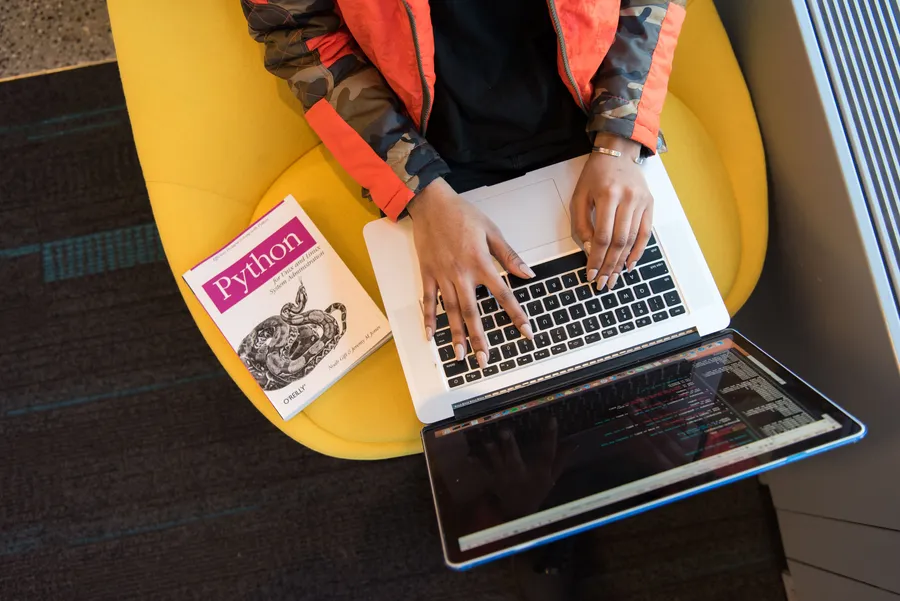 Person using a laptop displaying programming text beside a book