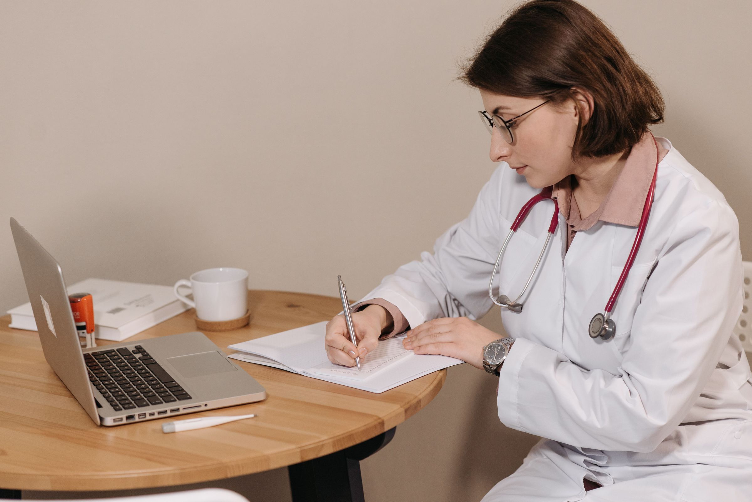 Doctor writing on paper at a desk beside a laptop