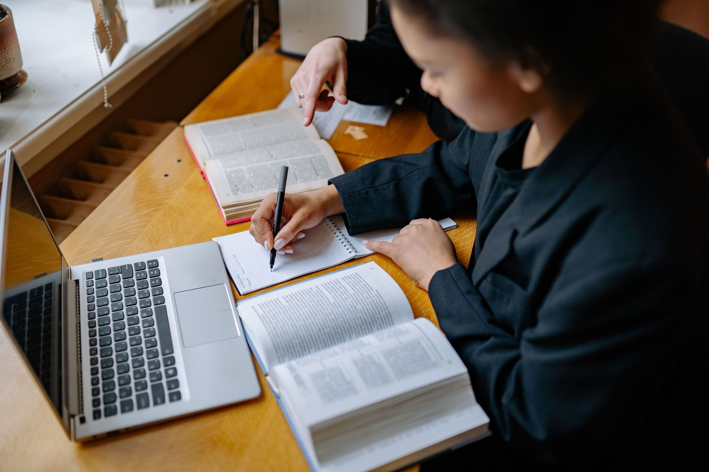 Person taking notebook notes while a colleague points to a book beside a laptop