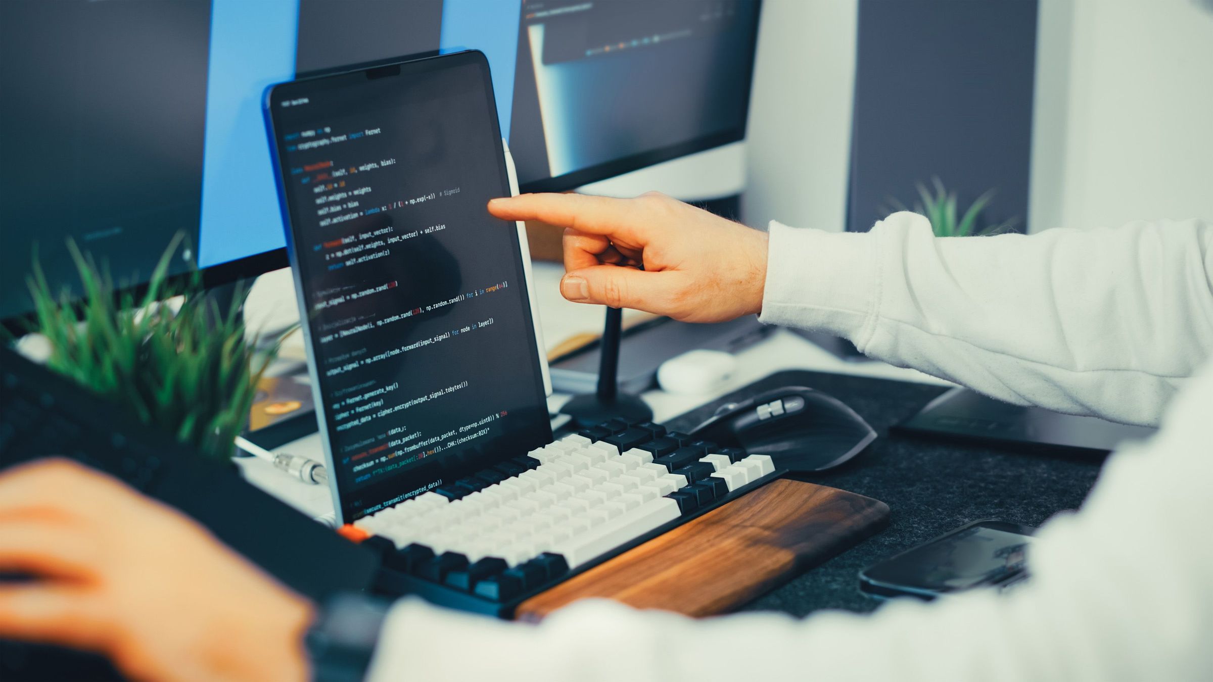 Hand pointing at code on a screen beside a keyboard and another monitor