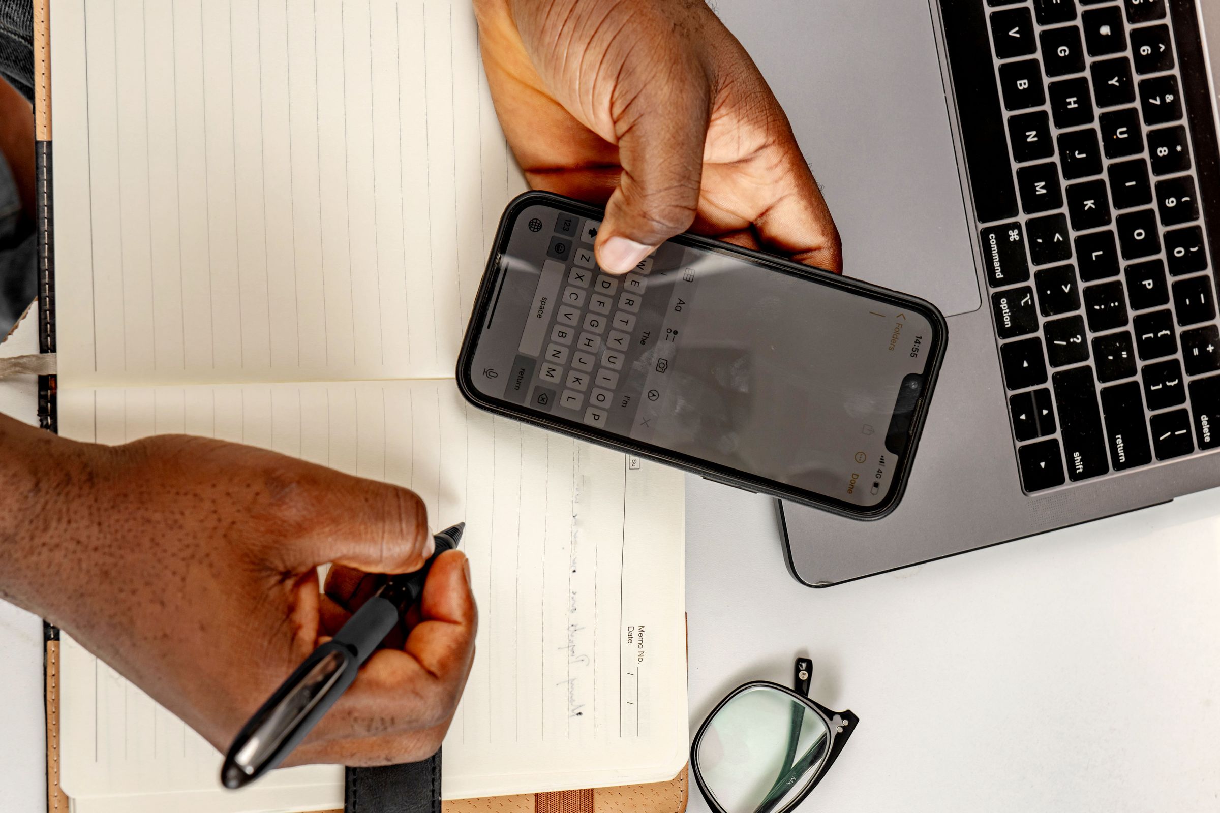 Hands using a phone and pen over a notebook beside a laptop
