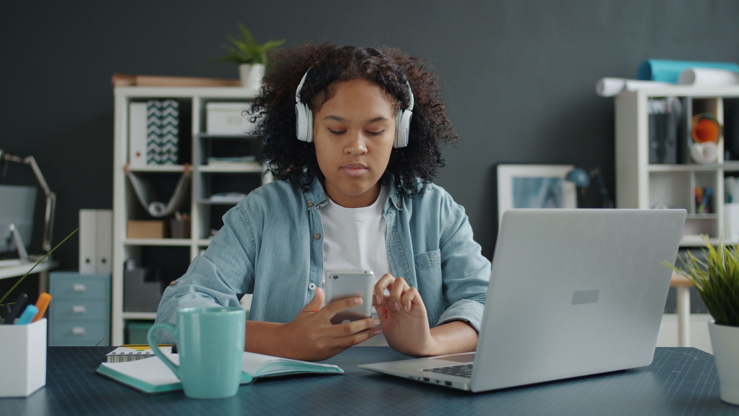 Woman checking a phone beside a laptop while wearing headphones