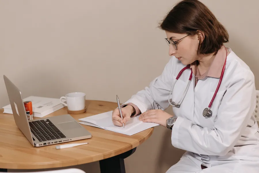 Doctor writing on paper at a desk beside a laptop