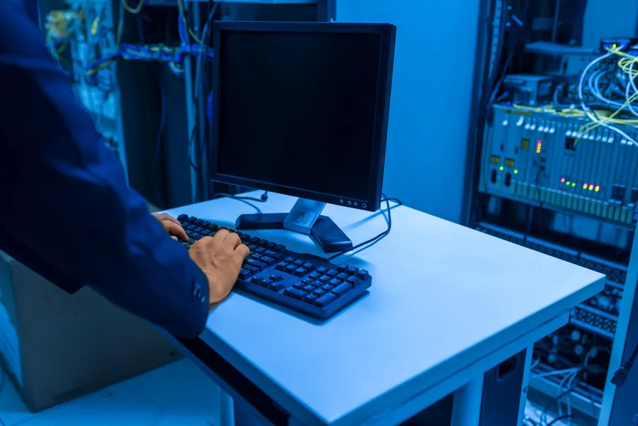 Technician using a keyboard beside server racks and a dark monitor