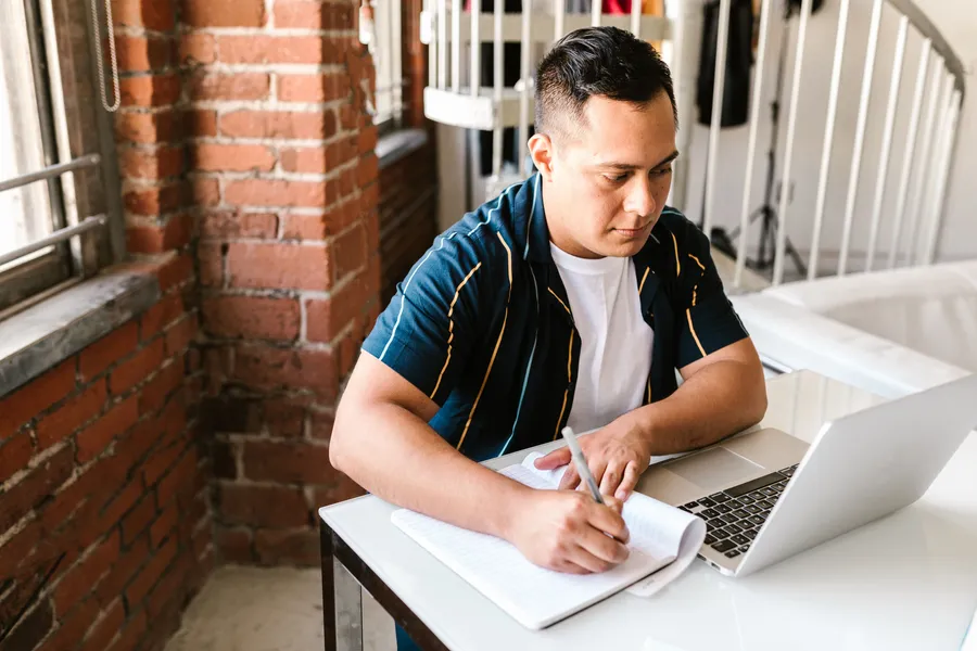 Man writing in a notebook in front of a laptop at a white table