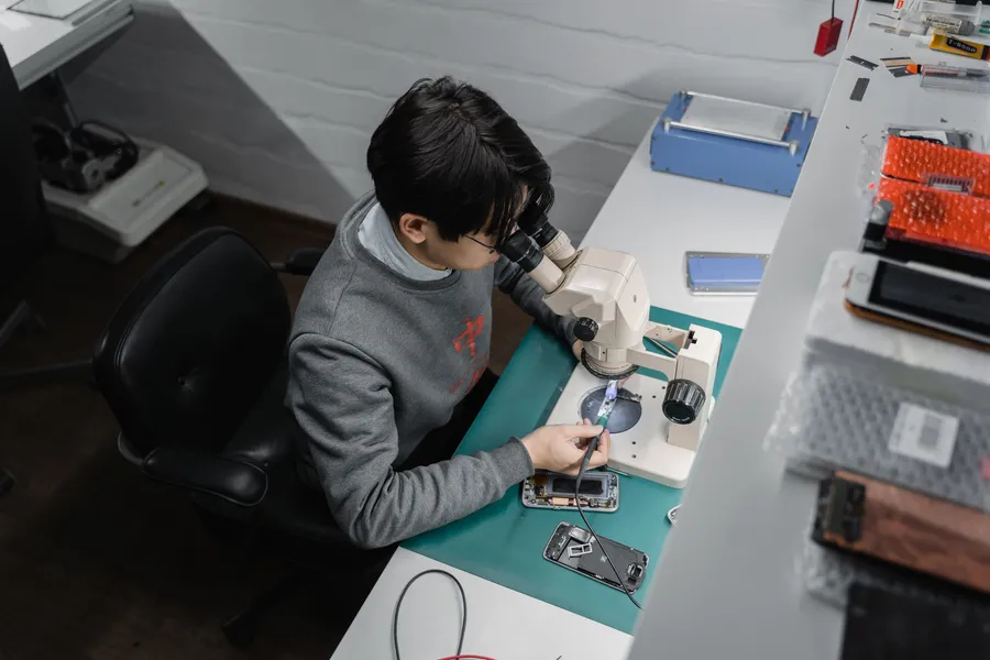 Technician working on phone components under a microscope at an electronics bench