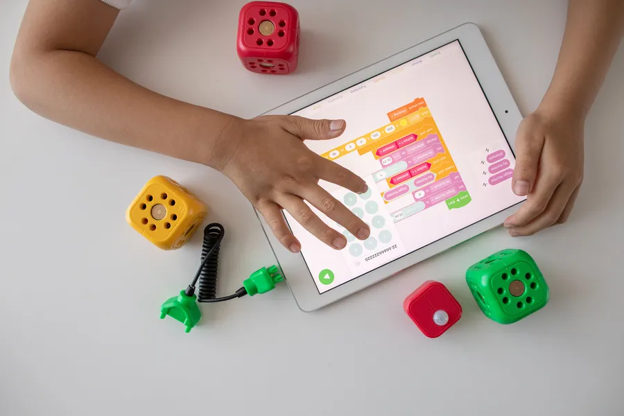 Child hands using a tablet displaying coding blocks beside educational cubes