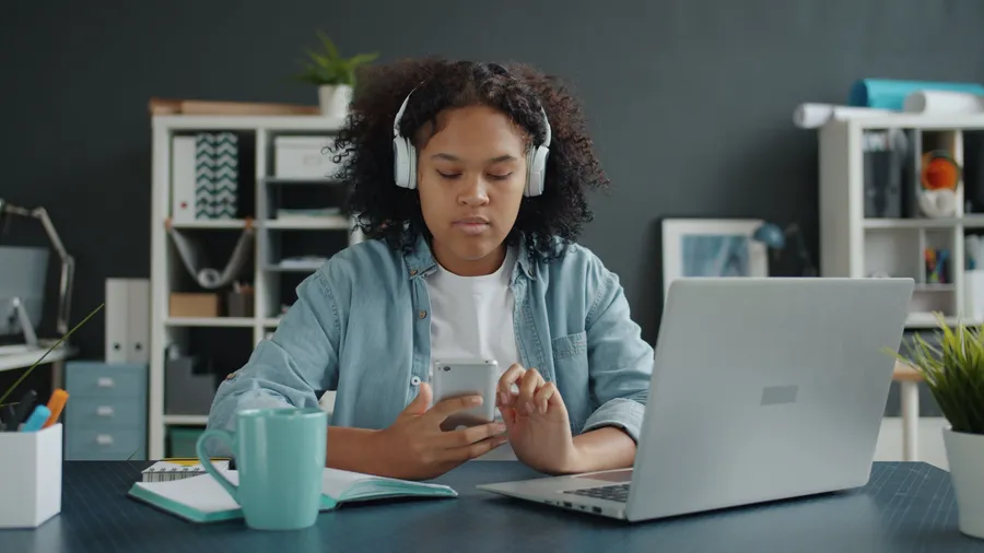 Woman checking a phone beside a laptop while wearing headphones